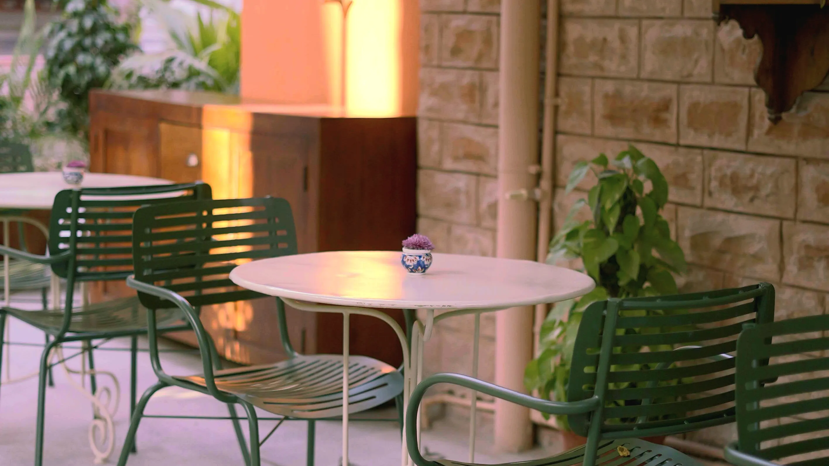 The open sandstone courtyard at Saanjh The Bistro in Ratanada, Jodhpur, with green iron chairs around pale stone tables and a brass lamp glowing against a terracotta wall at dusk