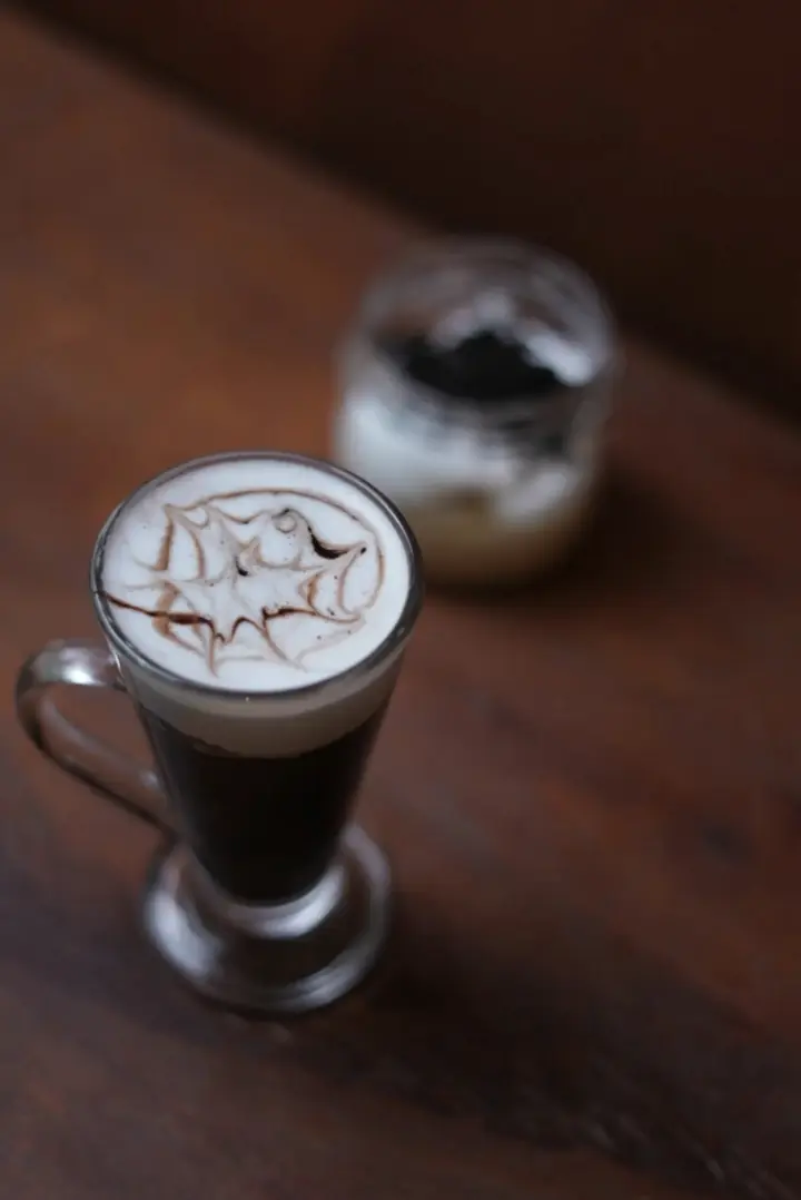 A tall glass of hot chocolate topped with whipped cream and a cocoa pattern, turning slowly on a wooden table at Saanjh The Bistro, Jodhpur